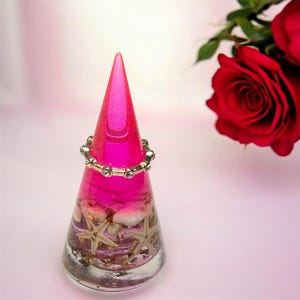 May include: A conical ring holder made of translucent hot pink resin, featuring embedded white starfish and gold-coloured flakes at the base. A silver-toned metal ring with small clear stones is placed around the centre of the cone.