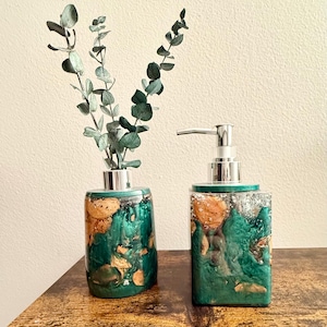 Op de afbeelding: Een set van twee groene en gouden hars zeepdispensers met zilveren pompen. De dispensers hebben een marmer effect en staan op een houten oppervlak.