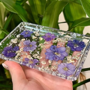 May include: A rectangular, clear resin tray with embedded purple flowers and silver flakes. The flowers are a deep violet hue, and the tray is held in a hand against a backdrop of green foliage. The tray is approximately 4 inches long and 3 inches wide.
