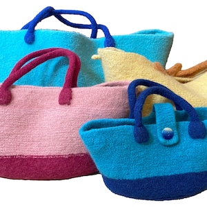 May include: Four knitted tote bags in different colours and sizes. The largest bag is light blue, the next largest is pink with a burgundy bottom, the third largest is yellow, and the smallest is blue with a navy bottom.