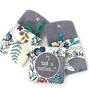 May include: Three small gray fabric pouches with floral patterns. The pouches have a flap that closes with a snap. One pouch has a measuring tape inside, another has a safety pin and a needle, and the third has a gray circle with the text "I had a notion..." embroidered on it.