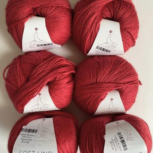 May include: Six balls of red Soft Lino Natural Line yarn are arranged on a white surface. Each ball has a label with the brand name and product details. The yarn is a vibrant, solid red color, perfect for knitting or crochet projects.