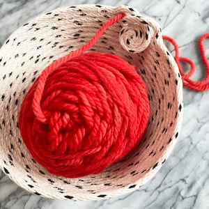 May include: A cream-coloured woven basket with black dotted detailing, containing a bright red ball of yarn. A length of red yarn trails from the ball, forming a decorative loop.