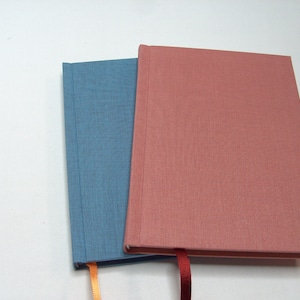May include: Two hardcover journals with fabric covers. One is blue and the other is pink. Both have ribbon bookmarks.