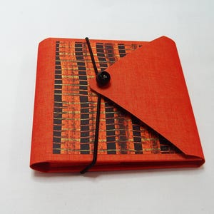 May include: A square, orange journal with a black elastic closure and a black button. The cover features a geometric pattern in black and gold. The journal's flap is folded over, revealing the interior.