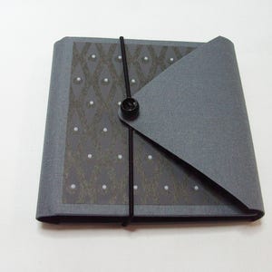 May include: A gray, square journal with a patterned cover featuring white dots and a black elastic closure. The journal has a flap that folds over and is secured with a black button.