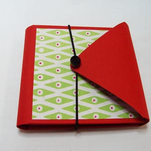 May include: A handmade red and patterned journal with a black elastic closure. The journal features a green and white patterned interior with red accents. The cover is a vibrant red, and the journal is secured with a black elastic band.