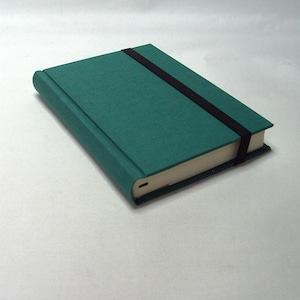 Könnte beinhalten: Ein teal-farbenes Notizbuch mit einem schwarzen Gummibandverschluss. Das Notizbuch hat einen strukturierten Einband und cremefarbene Seiten. Der Rücken ist abgerundet und das Notizbuch ist geschlossen. Das Notizbuch liegt auf einer weißen Oberfläche.