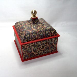 May include: A decorative box with a patterned lid and sides. The box features a red trim and a gold-colored finial on top. The patterned design includes shades of blue, green, and gold on a dark background. The box is a square shape.