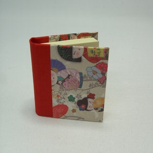 Op de afbeelding: Een klein, rood gebonden boek met een stapel plakbriefjes erin. De omslag heeft een kleurrijk patroon van Japanse poppen en bloemen.