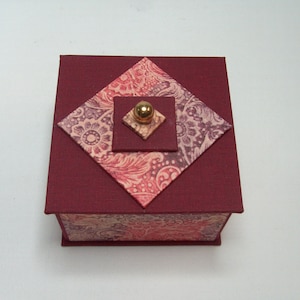 May include: A small, square, burgundy box with a patterned lid. The lid is made of four pieces of patterned paper, each with a different design. The box has a gold knob on the top.