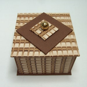 May include: A square box with a brown and beige checkered pattern. The box has a brown lid with a gold knob in the center.