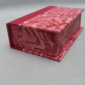 Könnte beinhalten: Eine kleine, rechteckige Schachtel mit einem roten Stoffbezug und einem gemusterten Design. Die Schachtel ist geschlossen und der Deckel ist leicht geöffnet.
