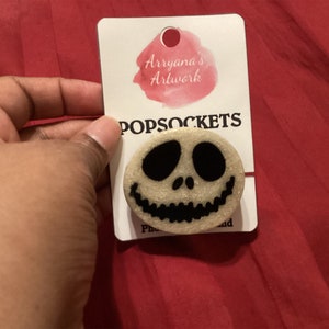 May include: A white and black popsocket with a glittery finish featuring a Jack Skellington face from the Nightmare Before Christmas movie. The popsocket is on a white card with the text "Arryana's Artwork" and "POPSOCKETS".