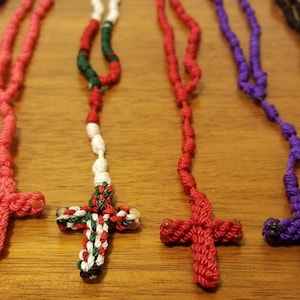 May include: Six knotted rope rosaries with different colored crosses. The rosaries are blue, red, white and green, red, purple, and black.