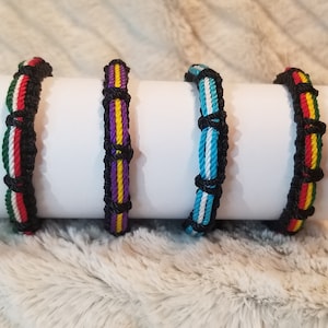 May include: Four colorful friendship bracelets with black string woven into each bracelet. The bracelets are displayed on a white surface.