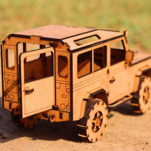 Peut inclure: Maquette en bois d'un SUV à quatre portes, avec une porte ouverte. Le modèle est en bois brun clair et présente des découpes détaillées pour les fenêtres, les roues et d'autres éléments. Le design rappelle un véhicule tout-terrain classique.