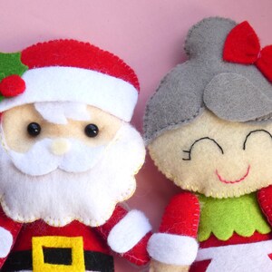 Puede incluir: Dos figuras de fieltro, una de Santa Claus con gorro rojo, barba blanca y adorno de acebo, y otra con cabello gris, lazo rojo y blusa verde. Ambas figuras presentan detalles faciales cosidos y est&aacute;n confeccionadas con capas de fieltro.