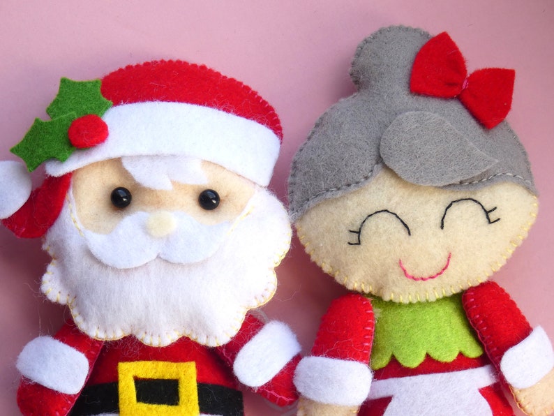 Puede incluir: Dos figuras de fieltro, una de Santa Claus con gorro rojo, barba blanca y adorno de acebo, y otra con cabello gris, lazo rojo y blusa verde. Ambas figuras presentan detalles faciales cosidos y est&aacute;n confeccionadas con capas de fieltro.