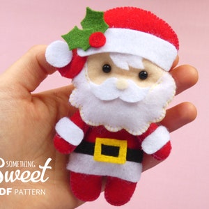 Puede incluir: Un adorno de fieltro de Santa Claus con traje rojo, barba blanca, cintur&oacute;n negro con hebilla amarilla y un detalle de hoja de acebo verde en el gorro. El texto "something Sweet PDF PATTERN" aparece impreso en blanco en la esquina inferior izquierda.
