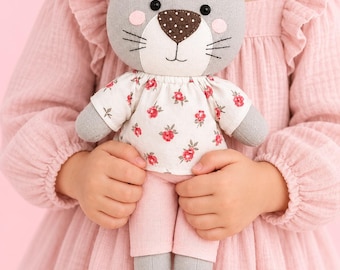 May include: A plush cat doll with gray fabric, a pink bow, black button eyes, and a brown polka-dot nose. The doll wears a white shirt with a red floral pattern and solid pink pants. The text SEWING PATTERN is printed at the bottom of the image.