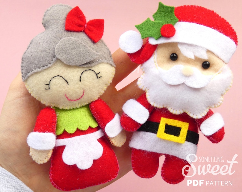 Puede incluir: Dos adornos de fieltro, una figura con mo&ntilde;o gris y lazo rojo, y Santa Claus con barba blanca y gorro de acebo. Ambas figuras tienen rasgos faciales cosidos y est&aacute;n vestidas con fieltro rojo, blanco, verde y negro con detalles de cintur&oacute;n amarillo.