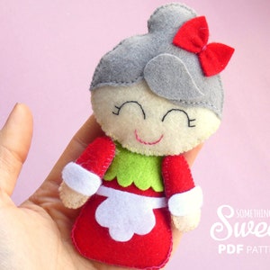 Puede incluir: Una mu&ntilde;eca de fieltro con un mo&ntilde;o gris y un lazo rojo, cara de color crema con ojos y boca bordados, corpi&ntilde;o verde y falda roja con delantal blanco. El texto "Something Sweet PDF PATTERN" es visible en la esquina inferior derecha.