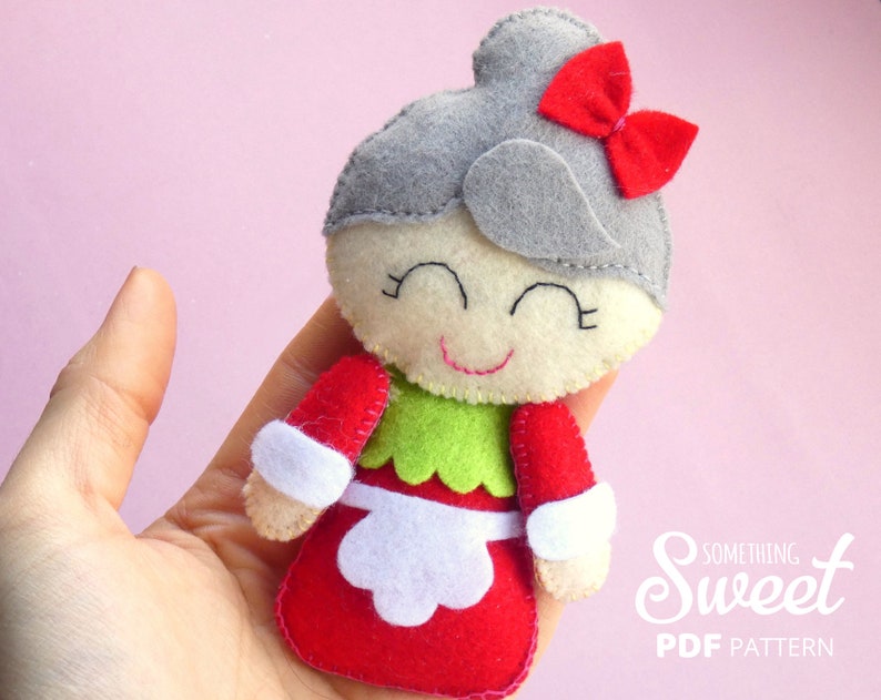 Puede incluir: Una mu&ntilde;eca de fieltro con un mo&ntilde;o gris y un lazo rojo, cara de color crema con ojos y boca bordados, corpi&ntilde;o verde y falda roja con delantal blanco. El texto "Something Sweet PDF PATTERN" es visible en la esquina inferior derecha.