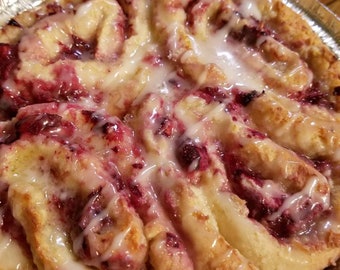 May include: A close-up of a homemade raspberry cinnamon roll with a white glaze. The roll is in a silver foil pan.