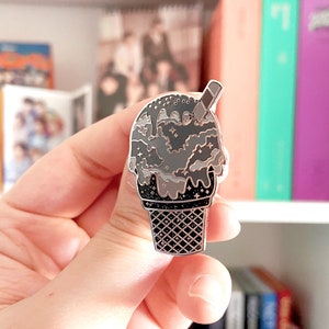 May include: A silver-colored enamel pin in the shape of an ice cream cone. The ice cream is a dark gray color with a chocolate drizzle and a small wafer stick. The cone is black with a waffle pattern.