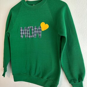 May include: A green sweatshirt with the word "MOM" appliqued in a plaid pattern, and a yellow heart. The sweatshirt has long sleeves and a crew neck. The garment is hanging on a wooden hanger.