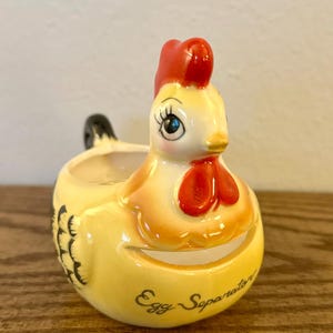 May include: A ceramic egg separator in the shape of a yellow hen with a red comb and wattle. The words "Egg Separator" are written on the front. The hen has black tail feathers and blue eyes.