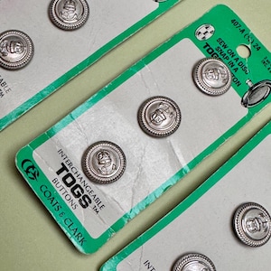 May include: Silver-toned interchangeable TOGS buttons on green and white packaging. The buttons feature nautical-themed designs and are from Coats & Clark. The packaging includes the text "SEW ON A DISC SNAP IN A TOG".