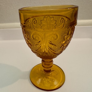 May include: An amber-colored glass goblet featuring a pressed pattern of embossed flowers and swirling scrollwork. The bowl of the glass has a textured background with a smooth rim and a sturdy stem leading to a circular base with floral details.