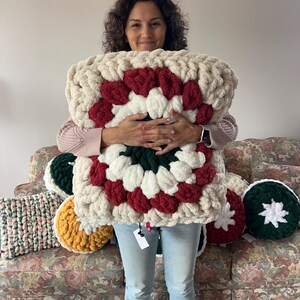 Pode incluir: Uma almofada grande, de malha grossa, com um design de quadrado da vov&oacute;. A almofada apresenta sec&ccedil;&otilde;es creme, vermelhas, brancas e verdes. Outras almofadas em v&aacute;rias cores s&atilde;o vis&iacute;veis no fundo, incluindo verde, amarelo e uma almofada listrada.