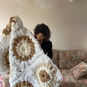 Pode incluir: Um cobertor grande, feito &agrave; m&atilde;o, de malha grossa, em tons de branco, creme e castanho. O cobertor apresenta um padr&atilde;o quadrado com desenhos circulares. A textura macia e felpuda &eacute; vis&iacute;vel. O cobertor est&aacute; sendo segurado em frente a um sof&aacute; com estampa floral.