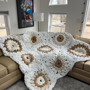 Pode incluir: Um cobertor de croch&ecirc; grosso, feito &agrave; m&atilde;o, em branco, creme e castanho. O cobertor apresenta um padr&atilde;o floral e est&aacute; drapeado sobre um sof&aacute; bege. O cobertor macio e texturizado adiciona um toque aconchegante &agrave; sala de estar.