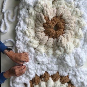 Pode incluir: Um close-up de um cobertor feito &agrave; m&atilde;o em andamento, apresentando um design quadrado com um padr&atilde;o central. O cobertor usa fios brancos, creme e marrons. Tesouras e fios soltos s&atilde;o vis&iacute;veis, indicando o processo de fabrica&ccedil;&atilde;o. O cobertor &eacute; macio e texturizado.