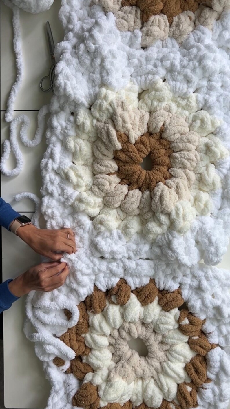 Pode incluir: Um close-up de um cobertor feito &agrave; m&atilde;o em andamento, apresentando um design quadrado com um padr&atilde;o central. O cobertor usa fios brancos, creme e marrons. Tesouras e fios soltos s&atilde;o vis&iacute;veis, indicando o processo de fabrica&ccedil;&atilde;o. O cobertor &eacute; macio e texturizado.