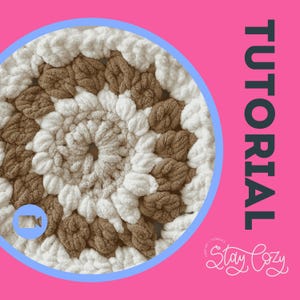Pode incluir: Um close-up de um item de croch&ecirc; circular feito &agrave; m&atilde;o em tons de branco e marrom. O padr&atilde;o de croch&ecirc; &eacute; vis&iacute;vel, com um design texturizado e em relevo. A palavra "TUTORIAL" &eacute; exibida verticalmente em um fundo rosa, junto com o texto "Stay Cozy".