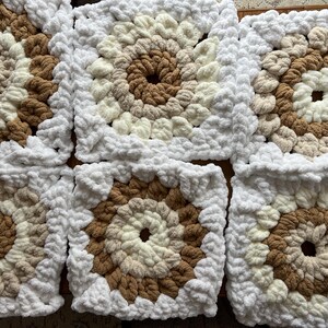 Pode incluir: Close-up de v&aacute;rios quadrados de croch&ecirc; em tons de marrom, creme e branco. Cada quadrado apresenta um c&iacute;rculo central marrom, rodeado por an&eacute;is conc&ecirc;ntricos de cores mais claras e uma borda branca. Os quadrados est&atilde;o juntos.