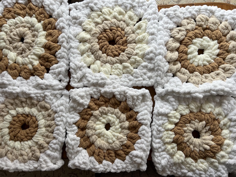 Pode incluir: Close-up de v&aacute;rios quadrados de croch&ecirc; em tons de marrom, creme e branco. Cada quadrado apresenta um c&iacute;rculo central marrom, rodeado por an&eacute;is conc&ecirc;ntricos de cores mais claras e uma borda branca. Os quadrados est&atilde;o juntos.