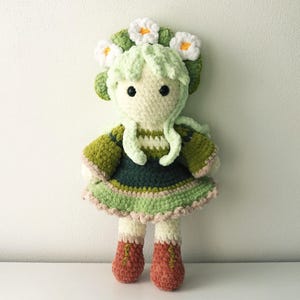 May include: A handmade crochet doll with a light green leafy crown and matching hair. The doll wears a green and dark green dress with a ruffled skirt and orange-brown boots. The doll is made of soft yarn.