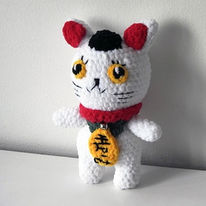 May include: White crocheted cat plush toy with red ears, black eyes, and a black nose. The cat is wearing a red and green scarf and a gold charm with the word "Happy" written on it.