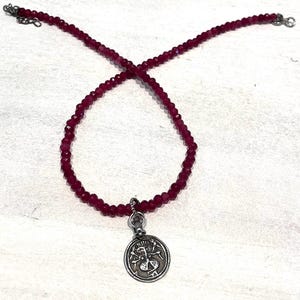 May include: A burgundy beaded necklace with a silver pendant. The pendant features a detailed design within a circular frame. The necklace is made of small, round beads and has a clasp closure.