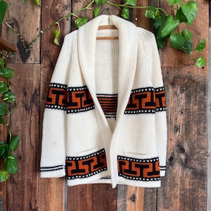 May include: Cream-colored cardigan sweater with a shawl collar and two front pockets. The sweater features geometric patterns in black and orange on the sleeves and lower hem. The sweater is hanging on a wooden hanger.