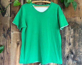 May include: Bright green t-shirt with a V-neck and short sleeves. The shirt has white trim on the neckline and sleeves. The shirt is hanging on a wooden hanger. The background is a wooden wall.