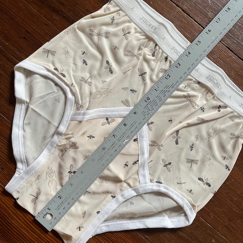Op de afbeelding: Twee beige slips met een libellenpatroon. De slips hebben een witte tailleband met de merknaam "Jockey" en witte randen. Een liniaal ligt over de slips en meet ongeveer 46 cm.