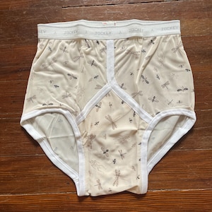 Op de afbeelding: Cr&egrave;mekleurig ondergoed met een libellenpatroon. De witte tailleband is bedrukt met het woord "Jockey". De randen zijn afgewerkt met witte biezen.