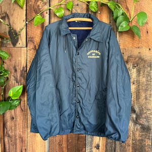 May include: Navy blue coach jacket with the text "University of Colorado" in gold lettering. The jacket has a button-up front, a drawstring at the bottom, and a dark blue fleece lining. The jacket is hanging on a wooden hanger.